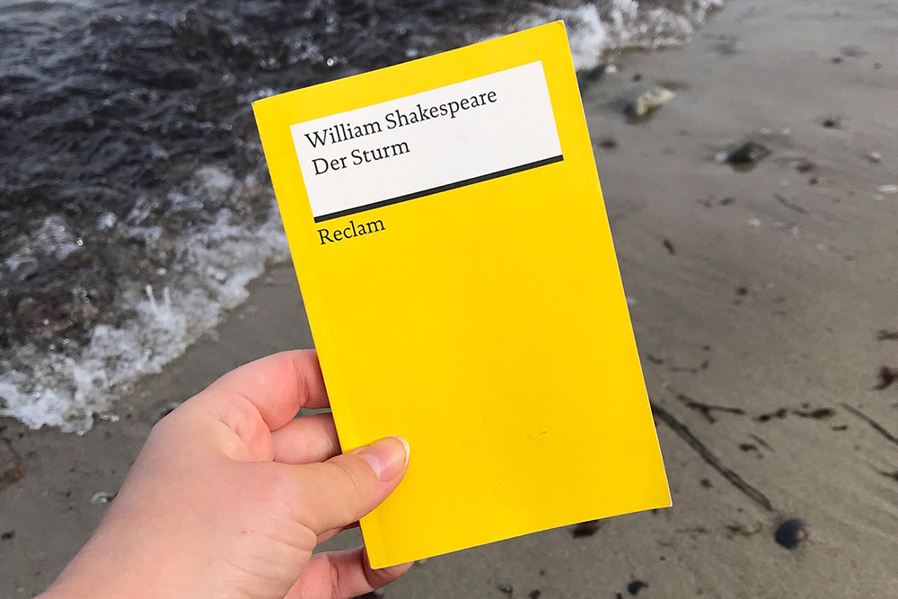 Eine Hand hält ein gelbes Reclambuch mit dem Titel "Der Sturm" von William Shakespeare. Im Hintergrund Strand und Meereswasser.